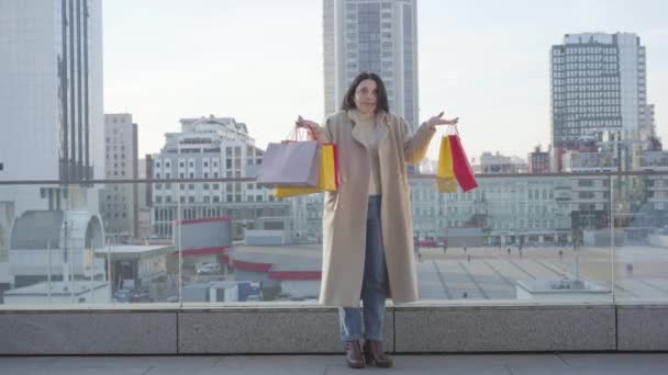 Mujer caucásica conmocionada levantando bolsas de compras coloridas y sonriendo. Feliz mujer adicta a las compras de pie en la calle de la ciudad con compras. Compras, estilo de vida, adicción . — Vídeos de Stock