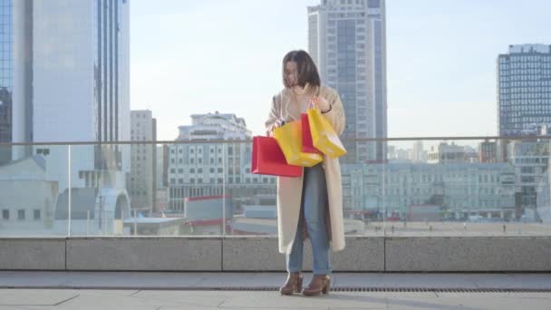 Happy Caucasian woman looking into her colorful shopping bags and smiling. Adult female shopaholic standing on city street with purchases. Shopaholism, lifestyle, addiction. — Stock Video