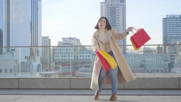 Retrato de una alegre mujer caucásica bailando con coloridas bolsas de compras y cantando. Feliz adicto a las compras disfrutando de compras en la calle de la ciudad. Compras, estilo de vida, adicción . — Vídeo de stock