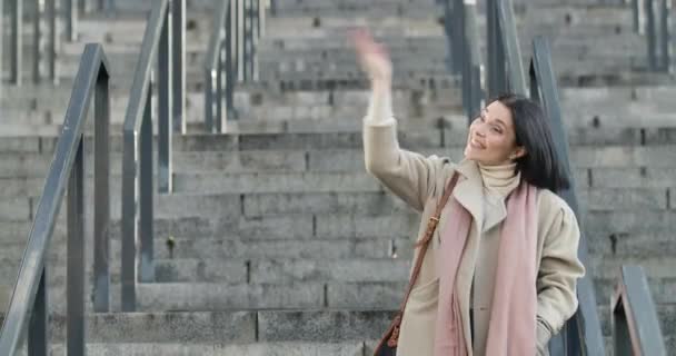 Belle femme brune caucasienne debout sur les escaliers en manteau élégant et agitant la main invitant quelqu'un. Jeune femme d'affaires positive faisant une pause dans la ville. Siège social Cinema 4k ProRes . — Video