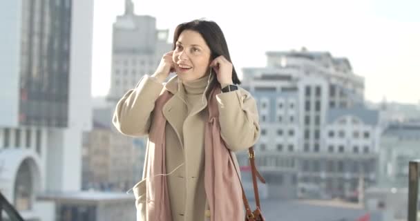 Portrait of charming Caucasian brunette woman putting on headphones and enjoying music outdoors. Positive elegant lady in beige coat resting in the city. Cinema 4k ProRes HQ. — Stock Video