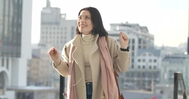 Прекрасна кавказька жінка в навушниках слухає музику на фоні хмарочосів. Виразна бізнесменка в бежевому пальто насолоджується проривом у місті. Фільм 4k Prores Hq. — стокове відео