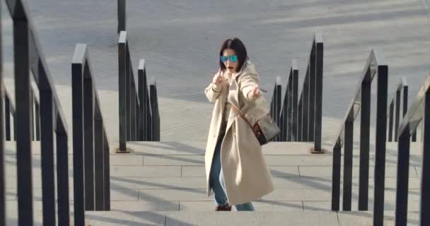 Portret van vrolijke volwassen blanke vrouw in zonnebril en beige jas dansen en zingen op straat. Brunette zakenvrouw draagt een koptelefoon en heeft plezier. Bioscoop 4k Prores Hq. — Stockvideo