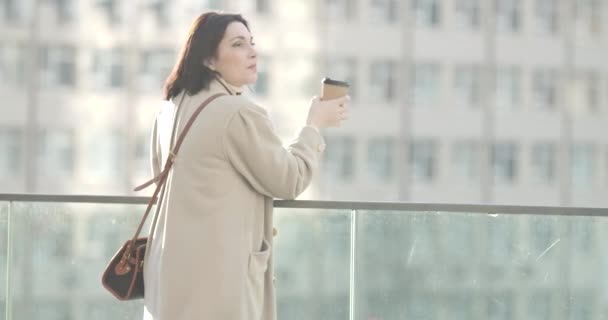 Vista trasera lateral de la joven mujer caucásica en elegante abrigo bebiendo café al aire libre en la ciudad. Exitosa empresaria confiada teniendo un descanso. Sede del cine 4k ProRes . — Vídeo de stock