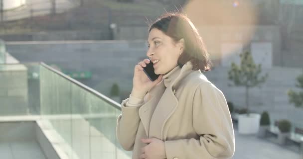 Zijaanzicht van een glimlachende blanke vrouw die in het zonlicht staat en aan de telefoon praat. Mooie brunette zakenvrouw die in de stad werkt. Bioscoop 4k Prores Hq. — Stockvideo