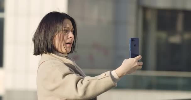 Vista lateral da mulher morena caucasiana fazendo rostos e tirando foto de selfie. Jovem positiva usando smartphone, se divertindo na rua da cidade. Cinema 4k ProRes HQ . — Vídeo de Stock