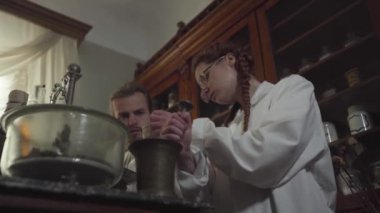 Kafkasyalı genç bir adam, kadın meslektaşına kuru yaprak veriyor. Kadın, havan ve tokmak kullanarak malzemeleri öğütüyor. Antik eczanedeki işçilerin portresi. Antika, 19. yüzyıl, tıp, sağlık hizmetleri..