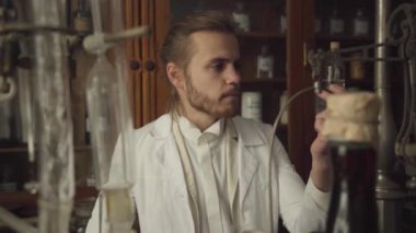 Beyaz cüppeli yakışıklı beyaz adam eski moda uyuşturucu şişesine bakıyor, konuşuyor ve genç kızıl saçlı kadına matara veriyor. Kamera test tüpünü takip ediyor. Antika eczane, retro, 19. yüzyıl.
