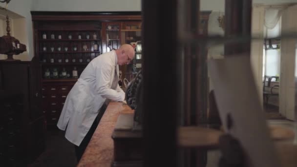 Widok z boku starego energicznego białego człowieka w białej szacie piórem. Aptekarz siedzi przy kasie i poprawia notatki. Apteka retro, apteka vintage, muzeum farmaceutyczne. — Wideo stockowe