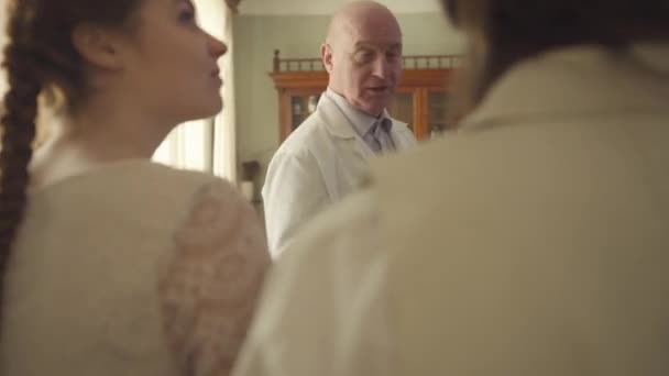 Tir sur l'épaule d'un jeune homme et d'une jeune femme caucasiens entrant dans une ancienne pharmacie. Apothicaire à tête chauve mature parlant aux visiteurs. Pharmacie vintage, style rétro, XIXe siècle . — Video