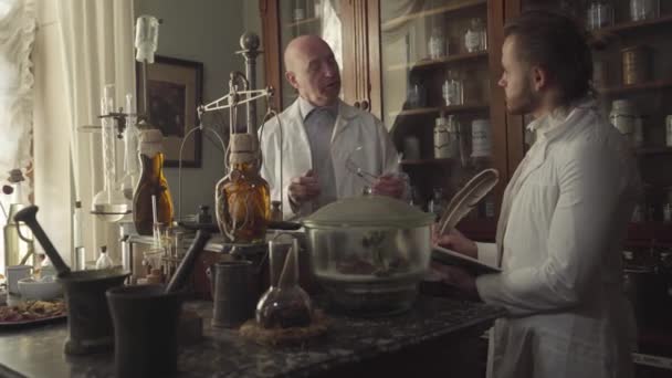 Viejo caucásico inteligente hablando con un joven nuevo empleado en una farmacia antigua. Profesional explicando el trabajo en farmacia. Retro farmacia, vintage, museo farmacéutico . — Vídeo de stock