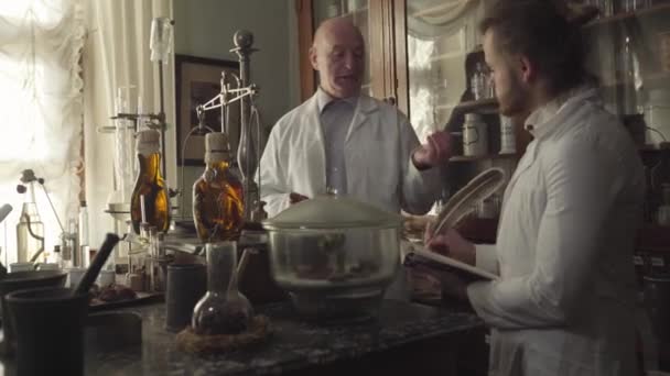 Två kaukasiska män står vid bordet med gamla kolvar, provrör och kemisk utrustning. Äldre farmaceuter som undervisar yngre kollega. Retroapotek. Kemiska termer skrivna på flaskor. — Stockvideo
