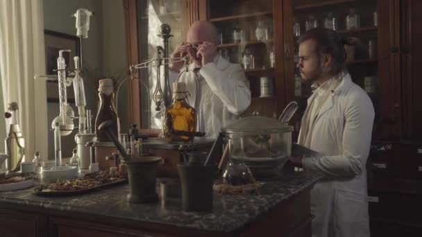 Old Caucasian pharmacist putting on eyeglasses and showing flask with transparent liquid to young man in white robe. New worker taking notes with feather pen. Chemical terms written on bottles. — Stock Video