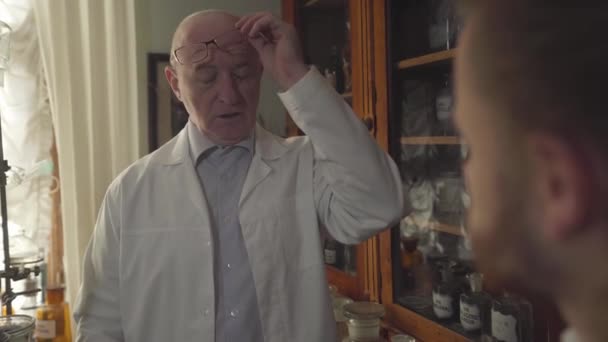 Portrait d'un homme âgé chauve en robe blanche enlevant des lunettes et parlant à un homme flou au premier plan. Ancien pharmacien caucasien éduquant un nouveau travailleur. Termes chimiques écrits sur bouteilles . — Video