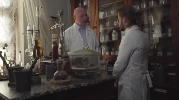 Portrait of bald Caucasian man yelling at younger worker and beating him with feather pen. Professional apothecary educating new employee in ancient drugstore. Chemical terms written on bottles. — Stock Video