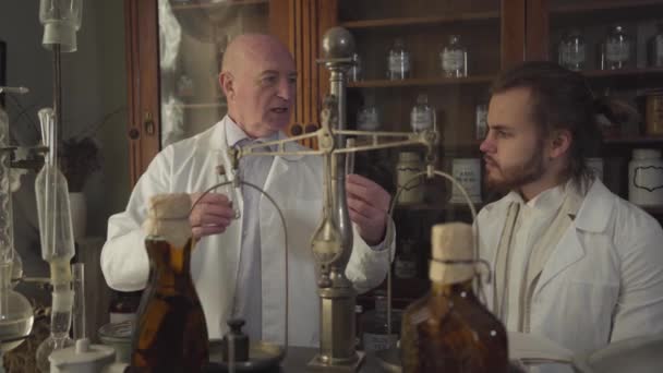 Feliz hombre caucásico viejo mostrando tubo de ensayo vintage y frasco para el hombre joven y hablando. Boticarios con túnicas blancas que trabajan en una farmacia antigua. Términos químicos escritos en botellas . — Vídeos de Stock