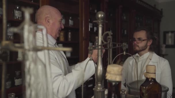 古代のフラスコとテストチューブを保持し、白いローブの若い同僚と話をする古いバルドヘッド薬剤師の側面図。2人の科学者が新薬を作った。ヴィンテージドラッグストア、 19世紀. — ストック動画