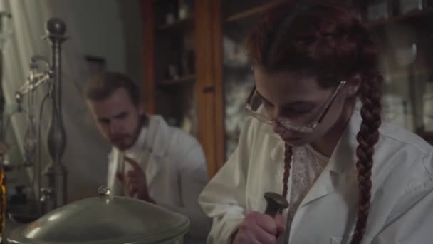 Close-up portrait of pretty redhead Caucasian girl with pigtails working in old-fashioned drugstore. Young woman in eyeglasses grinding medicaments with hands. Retro, vintage, pharmaceutical museum. — Stock Video
