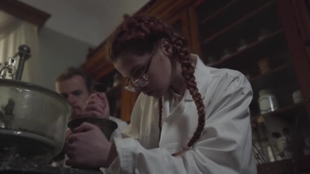 고대 약국에서 일하는 귀여운 코카서스 소녀의 맨 아래 사진. 젊은 빨간 머리 여자가 모르타르와 회전 병으로 재료를 갈고 있습니다. 뒤에 앉아 있는 남자 친구. — 비디오