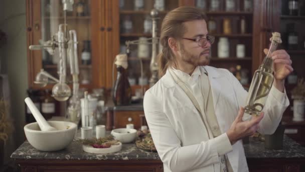 Молодой красивый белый мужчина в очках держит винтажную бутылку со змеей внутри, поворачивается к камере и улыбается. Фармацевт в белом халате стоит в древней аптеке. XIX век, винтаж . — стоковое видео