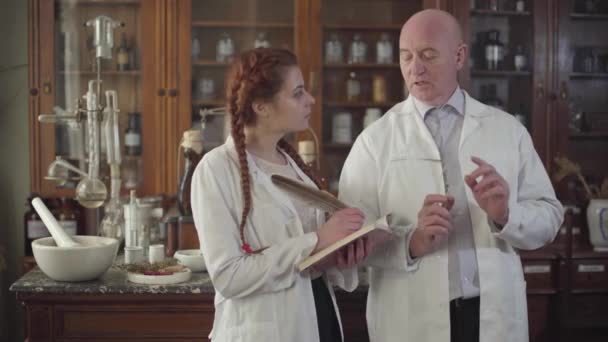 Belle fille rousse caucasienne avec des queues de cochon prenant des notes avec stylo plume. Homme âgé expliquant aux nouveaux pharmaciens les détails de leur travail dans une ancienne pharmacie. Pharmacie vintage, rétro, XIXe siècle . — Video