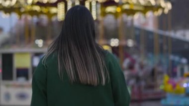 Kameralar beyaz bir kadının çocuk atlıkarıncalara yürüyüşünü gösteriyor. Sevimli kız kameraya dönüyor, gülümsüyor ve ileri doğru yürüyor. Neşeli bayan dışarıda dinleniyor. Yaşam tarzı, mutluluk, rahatlık.
