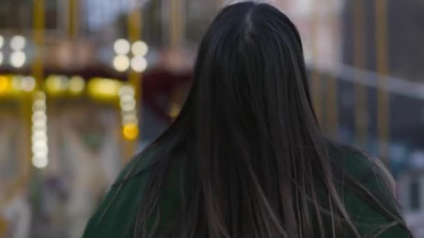 Back view of young pretty Caucasian woman walking to childrens carousels. Pretty girl turning to camera, smiling, and walking forward. Lifestyle, happiness, leisure. — ストック動画