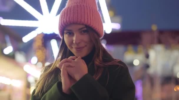 Portrait of charming Caucasian woman in pink hat looking at camera and smiling. Beautiful young girl spending evening on city market at the background of colorful carousels. Leisure, lifestyle. — ストック動画