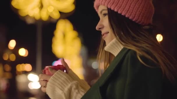 Vista lateral de la joven mujer caucásica de pie con taza de café al aire libre y mirando a su alrededor. Encantadora chica de sombrero rosa descansando en la noche en el mercado de la ciudad. Felicidad, ocio, estilo de vida . — Vídeo de stock