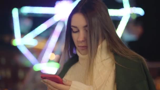 Portrait of cute Caucasian girl in white sweater using smartphone and smiling. Beautiful girl spending free time outdoors at the background of carousels. Lifestyle, leisure, communication. — ストック動画