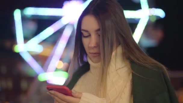 Retrato de linda chica caucásica en suéter blanco usando teléfono inteligente y sonriendo. Hermosa chica pasar tiempo libre al aire libre en el fondo de carruseles. Estilo de vida, ocio, comunicación . — Vídeos de Stock