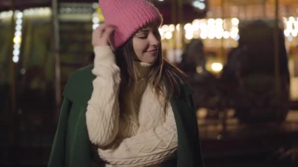 Χαμογελαστή λευκή κοπέλα με ροζ καπέλο και λευκό πουλόβερ να στέκεται έξω και να κοιτάζει τριγύρω. Όμορφη νεαρή γυναίκα που περνά το βράδυ στην αγορά της πόλης στο παρασκήνιο των πολύχρωμων καρουζέλ. — Αρχείο Βίντεο