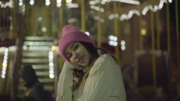 一个美丽的高加索女孩站在外面五颜六色的旋转木马前的画像。 穿着粉色帽子和白色毛衣的积极的年轻女性在城市市场上玩得很开心。 幸福、休闲、生活方式. — 图库视频影像