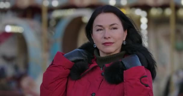 Close-up of middle-aged brunette Caucasian woman in red coat putting on hood. Beautiful positive lady standing on city street and waiting. Leisure, lifestyle, happiness. Cinema 4k ProRes HQ. — ストック動画