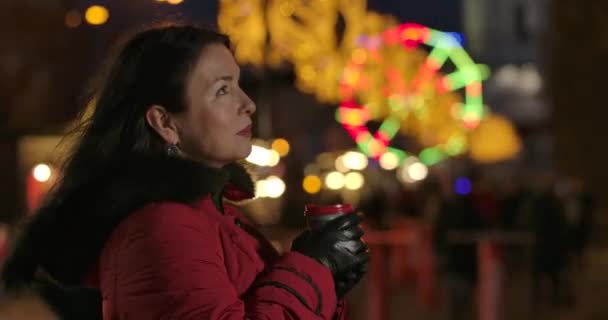 Side view of attractive adult Caucasian woman drinking coffee outdoors. Middle-aged brunette lady resting on city market at the background of colorful carousels. Cinema 4k ProRes HQ. — Stock Video