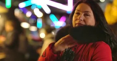 Kapüşonunu dışarıda tamir eden kırmızı ceketli beyaz bir kadın. Şehir pazarında renkli atlıkarıncaların arka planında duran büyüleyici kadının portresi. Sinema 4k Prores Merkezi.