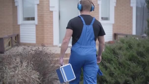 Vista trasera del hombre caucásico adulto en bata de trabajo azul y auriculares que llegan a la puerta de entrada. Fontanero con caja de herramientas que viene a realizar la reparación. Trabajo, profesional, ocupación . — Vídeo de stock