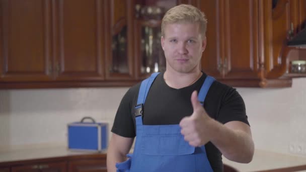 Portret van de jonge blanke klusjesman in een werkjas die in de keuken staat en duim omhoog laat zien. Knappe zelfverzekerde man tevreden met het resultaat van zijn werk. Professioneel beroep, garantiedienst. — Stockvideo