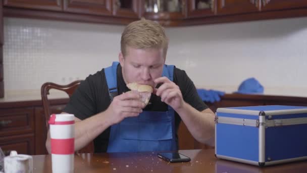 Portret młodego, białego hydraulika siedzącego w kuchni i jedzącego kanapki. Przystojniak dzwoni do kogoś przez smartfona. Odpoczynek, przerwa. — Wideo stockowe