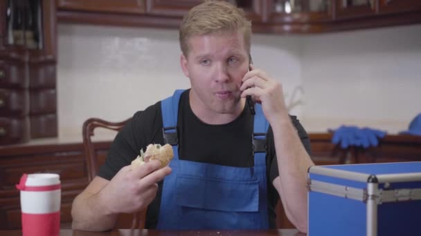 Portrait de jeune plombier caucasien parlant avec le client sur smartphone et manger un sandwich. Un rouquin qui fait une pause au travail. Repos, services professionnels . — Video