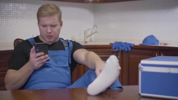 Porträtt av lat serviceman sitter i köket och använder smartphone. Vuxen vit man i blå arbetsrock vilar med benen på bordet. Arbete, vila, livsstil. — Stockvideo