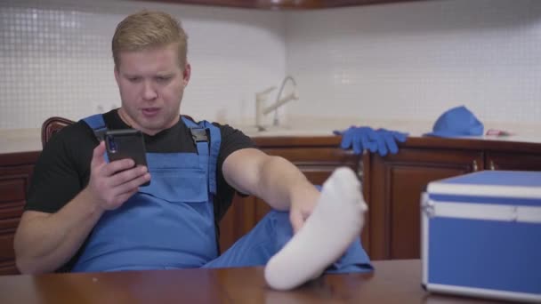 Retrato de un hombre de servicio perezoso sentado en la cocina y usando un teléfono inteligente. Hombre caucásico adulto en bata azul descansando con las piernas sobre la mesa. Trabajo, descanso, estilo de vida . — Vídeos de Stock