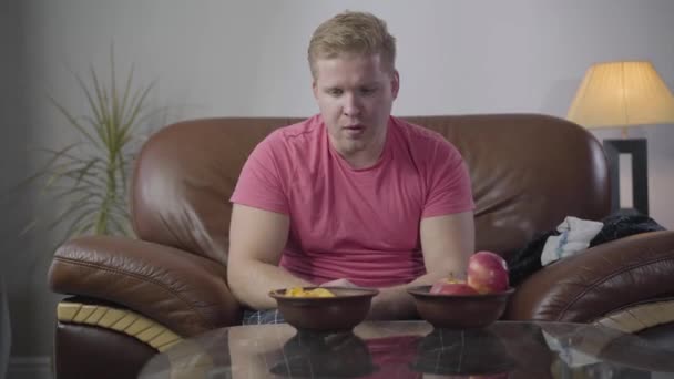 Portret van een jonge blanke man die op een koets zit en appels eet. Een mollige volwassen man met rood haar die alleen thuis rust. Lifestyle, eenzaamheid, ontspanning. — Stockvideo