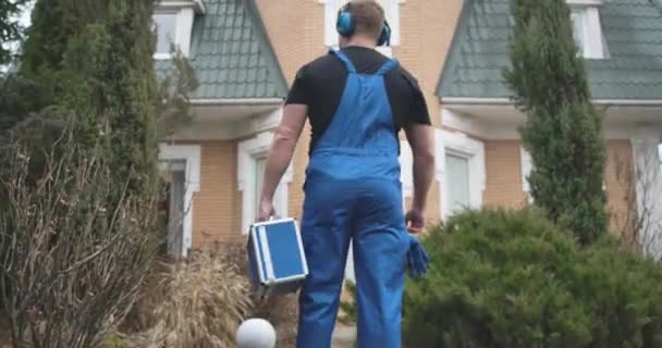 Vue arrière du jeune homme caucasien en écouteurs et peignoir bleu venant à la porte d'entrée de la maison. Travailleur avec boîte à outils venant effectuer la réparation. Travail, profession. Siège social Cinema 4k ProRes . — Video