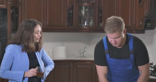 Retrato de la morena caucásica bebiendo té y hablando con el técnico en bata de trabajo. Pelirroja y joven dueño de la casa de pie en la cocina. Servicios profesionales de reparación. Sede del cine 4k ProRes . — Vídeos de Stock