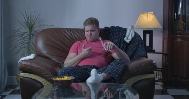 Portret grubego rudego białego mężczyzny siedzącego przed telewizorem i pijącego piwo. Samotny samotny facet spędzający wieczór sam w autokarze. Uzależnienie, samotność, alkoholizm. Kino 4k Prores Hq. — Wideo stockowe