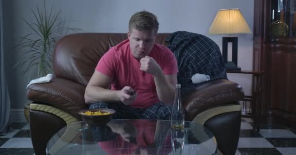 Portrait of indifferent Caucasian man sitting in front of TV and switching channels. Empty beer bottle standing on the table with chips. Loneliness, depression, lifestyle. Cinema 4k ProRes HQ. — Stock Video