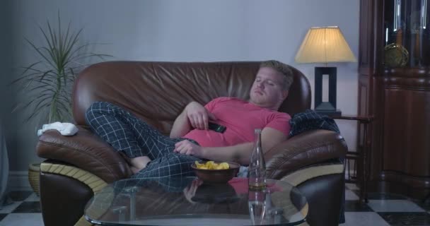 Lazy Caucasian man lying on sofa in front of TV and sleeping. Adult redhead guy resting with chips and empty beer bottle standing on the table. Lifestyle, addiction, alcoholism. Cinema 4k ProRes HQ. — 비디오