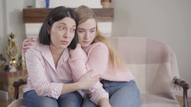 Portrait of young Caucasian brunette woman and blond girl sitting on couch at home and talking. Teenage daughter hugging mother. Family problems, unity, support. — Stock Video