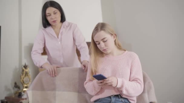 Portrait d'une jolie caucasienne blonde utilisant un smartphone et écoutant une femme adulte crier en arrière-plan. Adolescente se disputant avec sa mère à la maison. Incompréhension, conflit . — Video